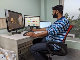 A person working at a white desk with three computer monitors. The person is seated on a red and black office chair and wearing a blue and white striped shirt and a black cap. The room has light green walls and a large window showing some greenery outside. Various cables and a white bag are visible under the desk.