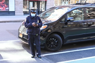 A uniformed officer wearing a mask is standing on the street near a black van. The officer appears to be holding a device, possibly a phone, and is looking towards the camera. The background includes a storefront and city street.