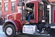 A red semi-truck parked on an urban street in front of a brick building. The driver's door is open, and a person in blue jeans and work boots is sitting with one foot on the step, appearing relaxed.