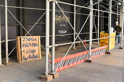 Outdoor scaffolding with metal poles surrounds a building under renovation. A wooden board displays the message 'It takes a child to raze a village. Paul Richard'. Nearby, a yellow construction dumpster is placed, along with an orange ladder leaning against the scaffold. A person wearing a hard hat and mask stands nearby, possibly a construction worker.