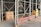 Outdoor scaffolding with metal poles surrounds a building under renovation. A wooden board displays the message 'It takes a child to raze a village. Paul Richard'. Nearby, a yellow construction dumpster is placed, along with an orange ladder leaning against the scaffold. A person wearing a hard hat and mask stands nearby, possibly a construction worker.