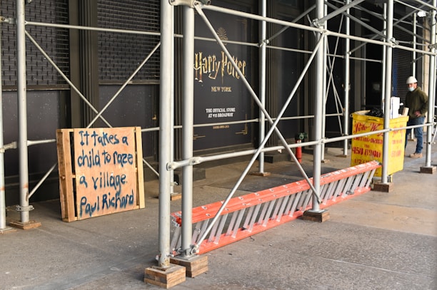 Outdoor scaffolding with metal poles surrounds a building under renovation. A wooden board displays the message 'It takes a child to raze a village. Paul Richard'. Nearby, a yellow construction dumpster is placed, along with an orange ladder leaning against the scaffold. A person wearing a hard hat and mask stands nearby, possibly a construction worker.