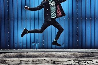 A person is mid-air in a dynamic jumping pose against a bright blue corrugated metal wall. The individual's lower half, wearing dark pants, is visible, with one leg raised higher than the other. The ground beneath is cracked and appears concrete.