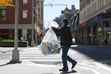 A person is carrying a large transparent bag filled with recyclable materials like plastic bottles and cans, walking across a city street crosswalk. The scene includes tall buildings lining the street, indicating an urban setting. The person is wearing casual clothing and a face mask, and the sun casts sharp shadows on the ground.