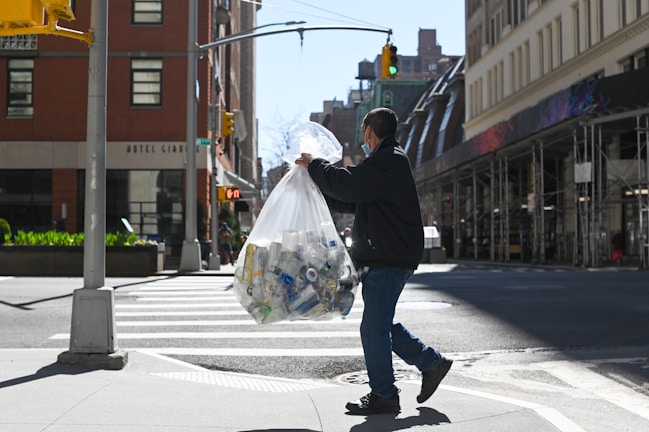 A person is carrying a large transparent bag filled with recyclable materials like plastic bottles and cans, walking across a city street crosswalk. The scene includes tall buildings lining the street, indicating an urban setting. The person is wearing casual clothing and a face mask, and the sun casts sharp shadows on the ground.