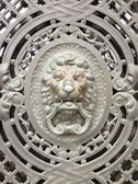 An intricately designed metal gate featuring an ornamental lion's head at the center. The lion's face is surrounded by elaborate scrollwork and geometric patterns, all crafted in a metallic finish. The artwork displays a high level of detail with a three-dimensional appearance, enhancing the regal and ornamental theme.