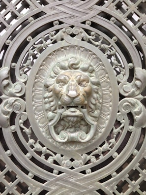 An intricately designed metal gate featuring an ornamental lion's head at the center. The lion's face is surrounded by elaborate scrollwork and geometric patterns, all crafted in a metallic finish. The artwork displays a high level of detail with a three-dimensional appearance, enhancing the regal and ornamental theme.