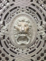 An intricately designed metal gate featuring an ornamental lion's head at the center. The lion's face is surrounded by elaborate scrollwork and geometric patterns, all crafted in a metallic finish. The artwork displays a high level of detail with a three-dimensional appearance, enhancing the regal and ornamental theme.
