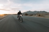 A person wearing a helmet rides a motorcycle down an open road with mountainous terrain in the background. The road appears to stretch out into the horizon, suggesting a sense of speed and freedom. The sun is setting or rising, casting a soft glow over the landscape.