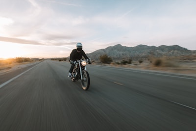 A person wearing a helmet rides a motorcycle down an open road with mountainous terrain in the background. The road appears to stretch out into the horizon, suggesting a sense of speed and freedom. The sun is setting or rising, casting a soft glow over the landscape.