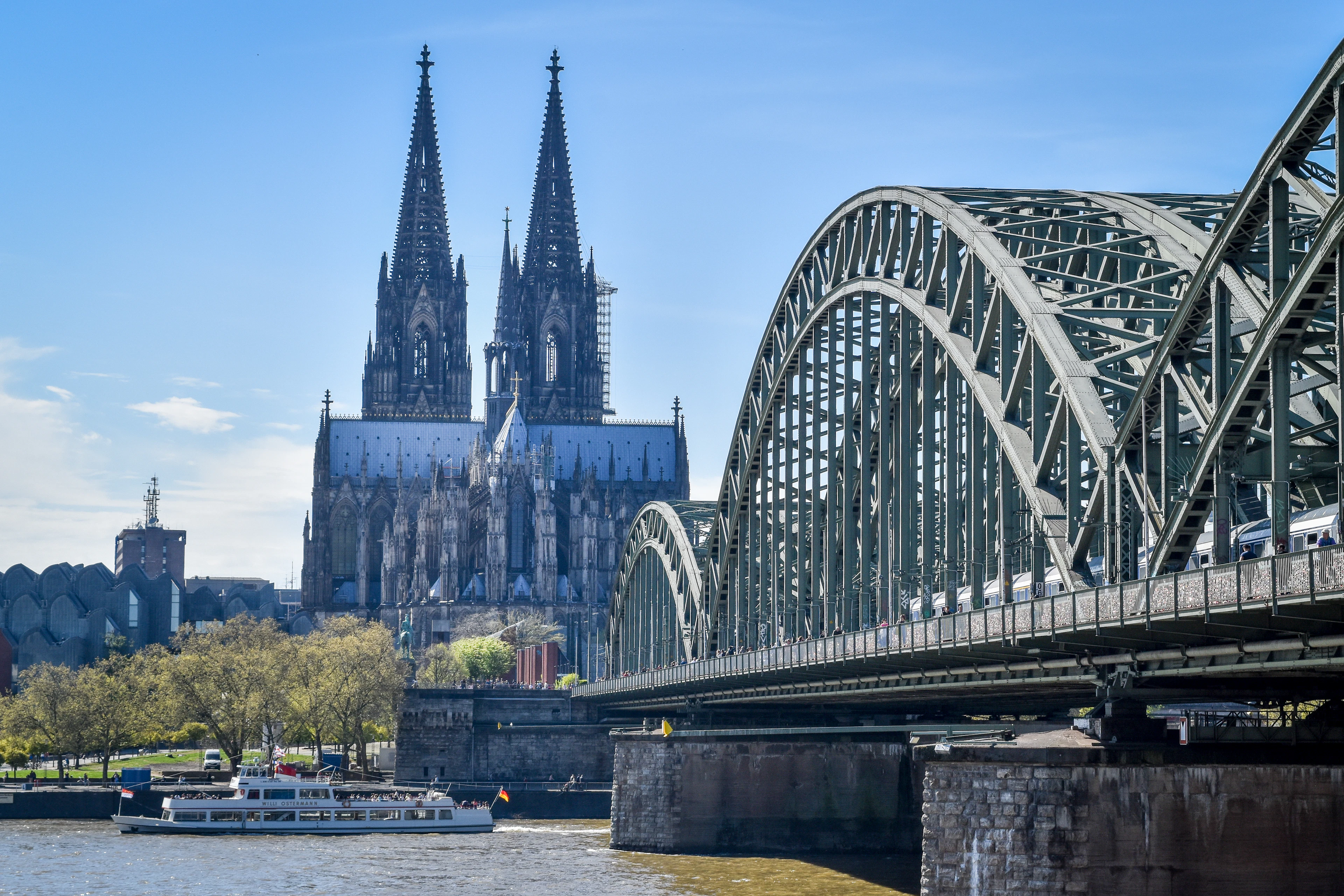 Cologne Cathedral rising beyond the steel arches of the Hohenzollern Bridge, with a boat passing on the Rhine