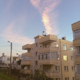 A series of apartment buildings with a boxy, modern design and pale beige exteriors. The sky is clear with pink-hued clouds stretching across it, bathed in warm light from the setting or rising sun. Telephone wires and a few trees are visible in the foreground, contributing to an urban residential setting.