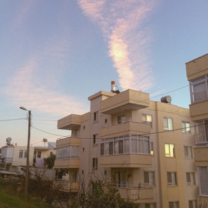 A series of apartment buildings with a boxy, modern design and pale beige exteriors. The sky is clear with pink-hued clouds stretching across it, bathed in warm light from the setting or rising sun. Telephone wires and a few trees are visible in the foreground, contributing to an urban residential setting.