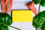 A bright yellow rectangular box with embossed lettering that reads 'MISS SWISS' is placed on a light wooden surface. Surrounding the box are large, vibrant green leaves and two reddish-orange tropical flowers, creating a fresh and lively aesthetic.