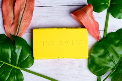 A bright yellow rectangular box with embossed lettering that reads 'MISS SWISS' is placed on a light wooden surface. Surrounding the box are large, vibrant green leaves and two reddish-orange tropical flowers, creating a fresh and lively aesthetic.