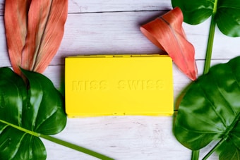 A bright yellow rectangular box with embossed lettering that reads 'MISS SWISS' is placed on a light wooden surface. Surrounding the box are large, vibrant green leaves and two reddish-orange tropical flowers, creating a fresh and lively aesthetic.