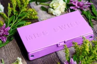A lavender-colored box with embossed text placed on a textured wooden surface, surrounded by lush green foliage and vibrant flowers in shades of purple, white, and yellow.
