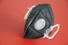 A black face mask with a filtration valve is positioned against a solid red background. The mask has white elastic straps and a textured material.