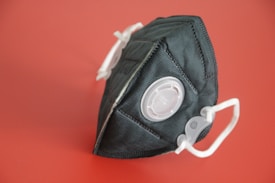 A black face mask with a filtration valve is positioned against a solid red background. The mask has white elastic straps and a textured material.