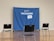 A blue banner with the word 'VACCINATED!' and a message encouraging COVID-19 vaccination is set up as a backdrop. It is surrounded by three black and gray chairs on a polished wooden floor in an empty room.