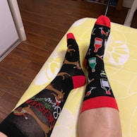Two legs are stretched out on a yellow-patterned bedspread, wearing mismatched socks. One sock features Christmas-themed reindeer designs, while the other displays bottles of alcohol and wine glasses. Both socks have red cuffs and toes. The surrounding environment includes a wooden floor and a portion of a room corner.