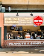 A concession stand with a sign advertising Polish sausage, jumbo refillable popcorn, and other snacks. The signage includes a Coors Field Fanfare logo. The stand is constructed with brick and features several menu boards and snack items.