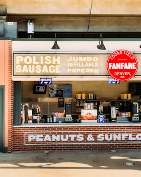 A concession stand with a sign advertising Polish sausage, jumbo refillable popcorn, and other snacks. The signage includes a Coors Field Fanfare logo. The stand is constructed with brick and features several menu boards and snack items.