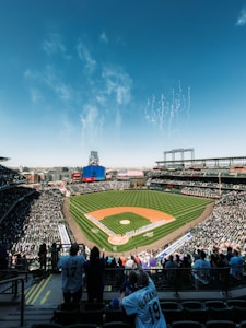A large baseball stadium filled with spectators, featuring a well-manicured green field with a pitcher's mound in the center. Spectators are standing and cheering, and a group of people in uniform are lined up on the field. Fireworks are visible in the blue sky above the stadium. There are jersey-clad fans visible in the foreground and large screens and billboards around the stadium.