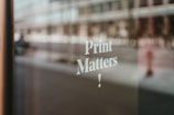 A blurred reflection of an urban street scene visible through a glass window with the words 'Print Matters!' printed in white, showcasing a focus on typography.