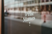 A blurred reflection of an urban street scene visible through a glass window with the words 'Print Matters!' printed in white, showcasing a focus on typography.