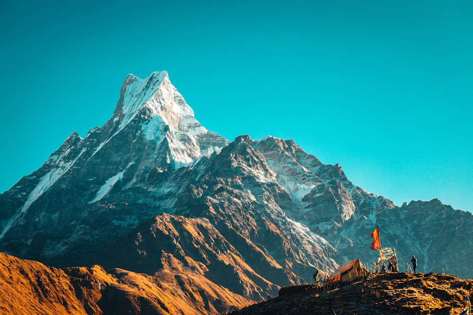 The image depicts a majestic mountain range with snow-capped peaks against a clear blue sky. The mountains, painted in shades of brown and gray, rise high into the sky, creating a sense of grandeur and scale. Atop one of these peaks is a small hut or structure, possibly used for camping or other outdoor activities. In the foreground, there are several people standing on the mountain's edge, likely hikers or adventurers exploring the area. The image conveys a sense of adventure and exploration, with the natural beauty of the mountains contrasting against the clear blue sky.