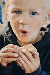 A young child with blue eyes and blonde hair is eating a messy s'more, with chocolate and marshmallow on their face and hands. The child is wearing a dark-colored hoodie and is focused on the treat.