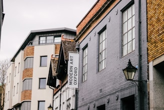 A street view featuring buildings with varied architectural styles. The structure on the left has a modern design with large windows and brick accents, while the building on the right is older, characterized by grey stonework and traditional window designs. A sign for Modern Art Oxford hangs on one of the buildings. Street lamps are visible, adding an element of classic style.