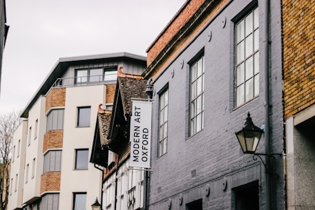 A street view featuring buildings with varied architectural styles. The structure on the left has a modern design with large windows and brick accents, while the building on the right is older, characterized by grey stonework and traditional window designs. A sign for Modern Art Oxford hangs on one of the buildings. Street lamps are visible, adding an element of classic style.