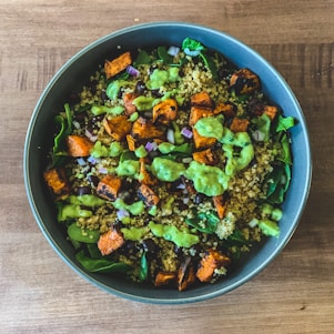 A bowl filled with a quinoa salad featuring roasted sweet potato chunks, fresh greens, black beans, chopped red onions, and drizzled with a green dressing.