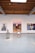 An art gallery with white walls displays three large, colorful paintings. A person wearing a white dress and neon green shoes stands in the middle of the room. The gallery has an industrial style ceiling with exposed wooden beams and spotlights.