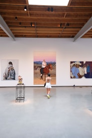An art gallery with white walls displays three large, colorful paintings. A person wearing a white dress and neon green shoes stands in the middle of the room. The gallery has an industrial style ceiling with exposed wooden beams and spotlights.
