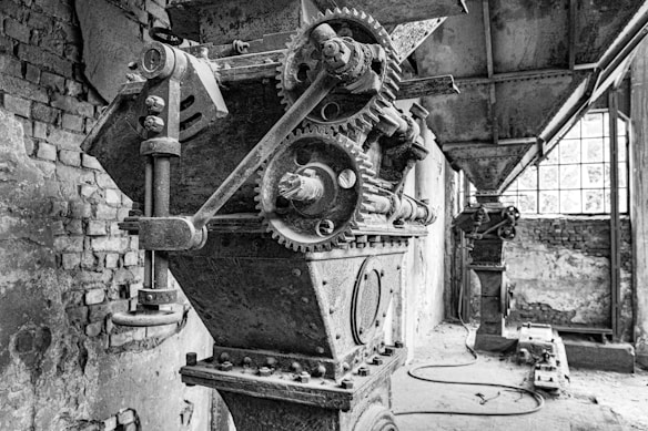 An industrial scene featuring large, mechanical machinery in an old and possibly abandoned factory setting. The image includes detailed gears, bolts, and a metal structure with a worn, rustic appearance. The background shows a brick wall, grime-covered floors, and large windows allowing some light to enter.