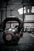 Closer shot of a worn kettlebell on a speckled gym floor, with muted lighting in a fitness facility. Background reveals additional gym equipment and an industrial-style setting.