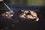 Several pieces of meat are cooking on a barbecue grill, with grill marks visible on the surface. Smoke rises from the grill, indicating that the meat is being cooked over an open flame. The environment suggests an outdoor setting with sunlight giving a warm glow to the scene.
