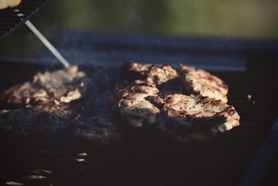 Several pieces of meat are cooking on a barbecue grill, with grill marks visible on the surface. Smoke rises from the grill, indicating that the meat is being cooked over an open flame. The environment suggests an outdoor setting with sunlight giving a warm glow to the scene.