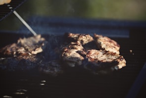 Several pieces of meat are cooking on a barbecue grill, with grill marks visible on the surface. Smoke rises from the grill, indicating that the meat is being cooked over an open flame. The environment suggests an outdoor setting with sunlight giving a warm glow to the scene.
