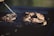 Several pieces of meat are cooking on a barbecue grill, with grill marks visible on the surface. Smoke rises from the grill, indicating that the meat is being cooked over an open flame. The environment suggests an outdoor setting with sunlight giving a warm glow to the scene.