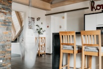 A modern kitchen area with wooden elements. There are two bar stools with cushions, a kitchen counter with a black surface, and a stone accent wall. A white hanging chair sways slightly, blurring the image slightly, and plants are placed on a wooden stand. Light-colored wood beams and ceiling slats create a warm ambiance.