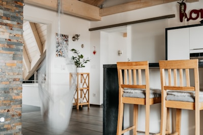 A modern kitchen area with wooden elements. There are two bar stools with cushions, a kitchen counter with a black surface, and a stone accent wall. A white hanging chair sways slightly, blurring the image slightly, and plants are placed on a wooden stand. Light-colored wood beams and ceiling slats create a warm ambiance.