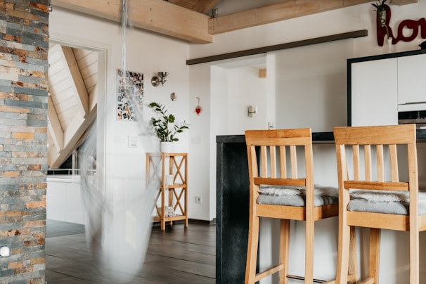 A modern kitchen area with wooden elements. There are two bar stools with cushions, a kitchen counter with a black surface, and a stone accent wall. A white hanging chair sways slightly, blurring the image slightly, and plants are placed on a wooden stand. Light-colored wood beams and ceiling slats create a warm ambiance.