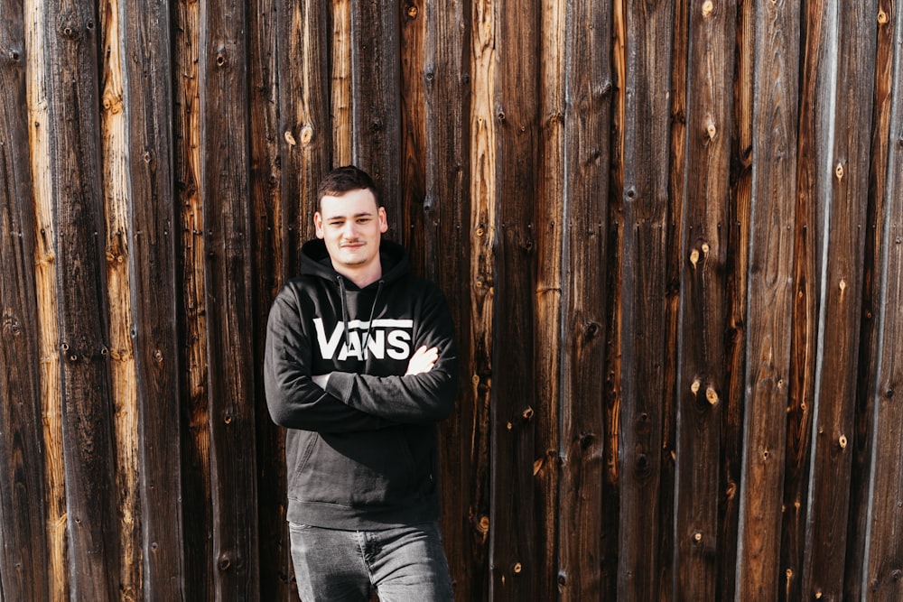 man in black and white hoodie and blue denim jeans leaning on brown wooden wall