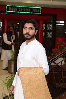 A man in a white traditional outfit is holding a woven fabric with intricate designs. The setting appears to be the entrance of a restaurant, indicated by a sign above that reads 'NANO CHINESE THAI'. In the background, another person is speaking on a phone.