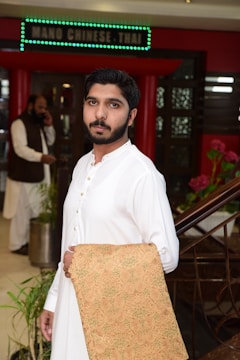 A man in a white traditional outfit is holding a woven fabric with intricate designs. The setting appears to be the entrance of a restaurant, indicated by a sign above that reads 'NANO CHINESE THAI'. In the background, another person is speaking on a phone.