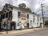 A two-story white building with several advertisements and colorful signs on the walls. It has a large sign that reads 'Drink Regal Beer.' The structure has multiple air conditioners, dark shutters, and posters displaying menu items like waffles and po'boys. There are plants in pots along the sidewalk and a street with power lines overhead.